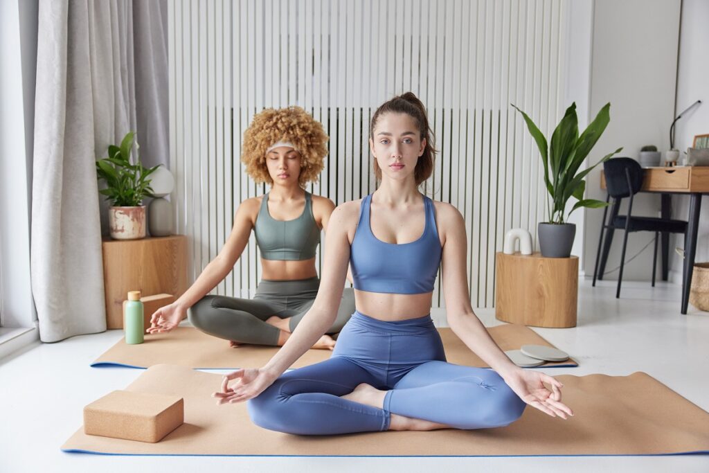 Yoga practice and meditation concept. Two women sit crossed legs on fitness mat breath deeply dressed in tracksuit try to relax after hard working day find balance pose against cozy interior