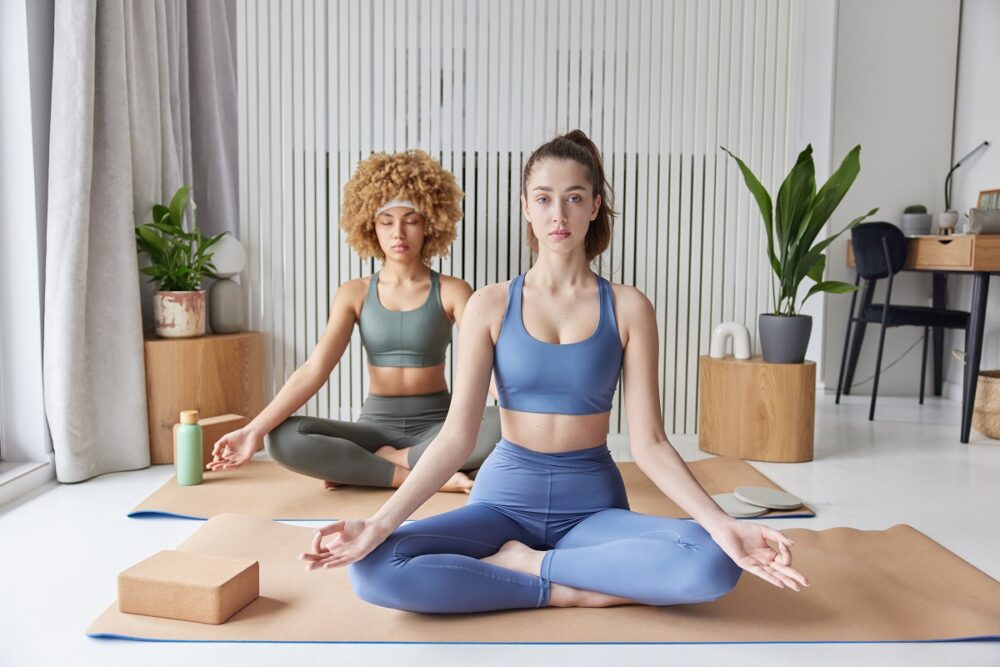 Yoga practice and meditation concept. Two women sit crossed legs on fitness mat breath deeply dressed in tracksuit try to relax after hard working day find balance pose against cozy interior