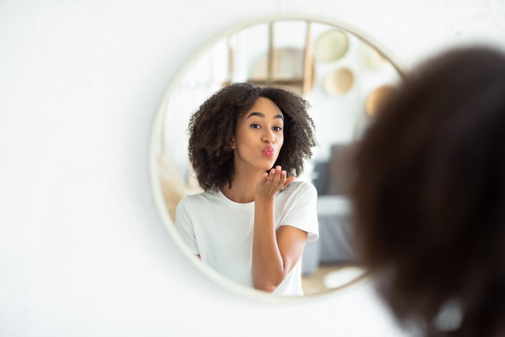Great appearance, love yourself and perfect skin. Cheerful young african american lady makes air kiss and looks in reflection of round mirror in living room, on white wall, panorama, free space