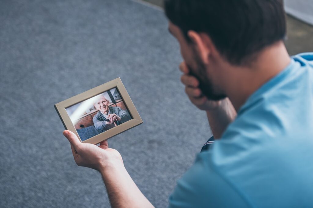 back view of adult man grieving while looking at photo frame with old man at home