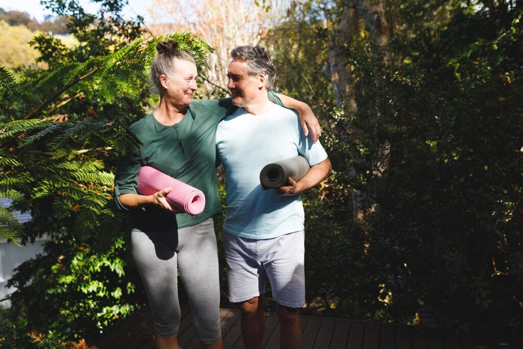 Happy senior caucasian couple practicing yoga, holding yoga mats in sunny garden. healthy retirement lifestyle, spending time at home. 