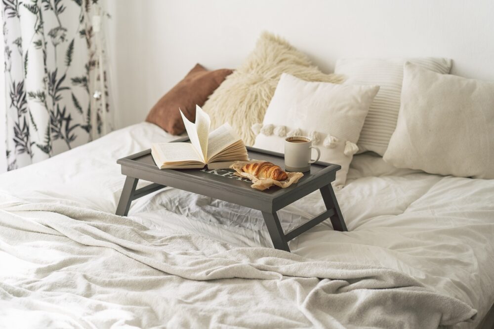 Tray with croissant, cup of coffee and book in white clean bed. Mindful and quiet living. Breakfast in Bed
