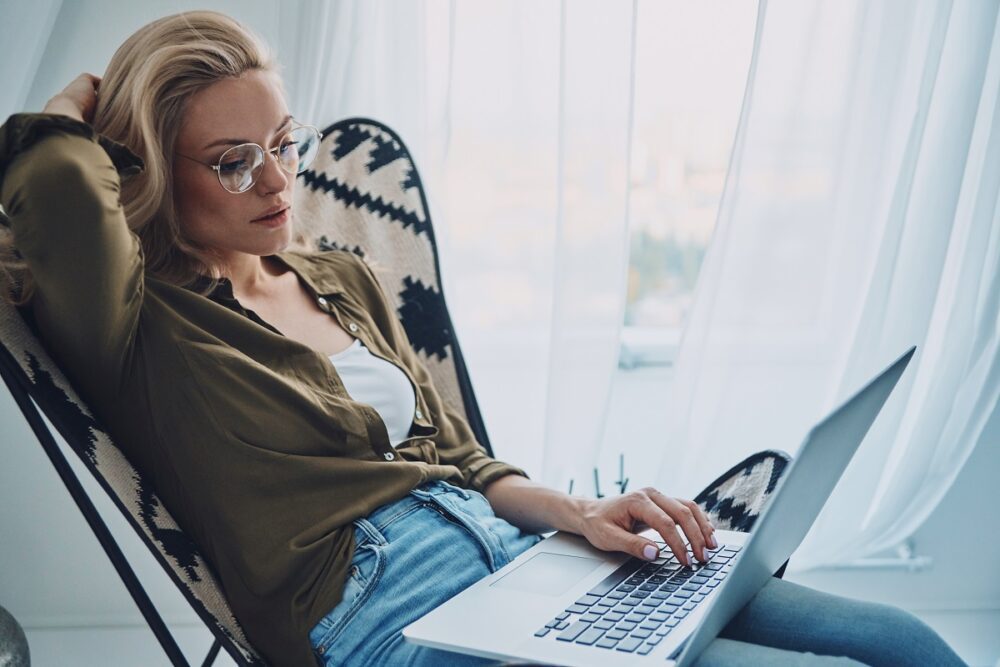 Attractive young woman using laptop while sitting in comfortable chair at home