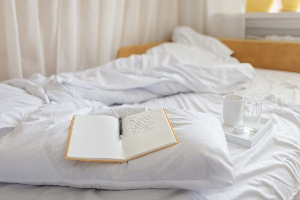 Mental health care, positive thinking and wellness concept. Woman lying in bed, practicing stress relief challenge with journal about feeling, glass of water and gratitude list, focus on word mental