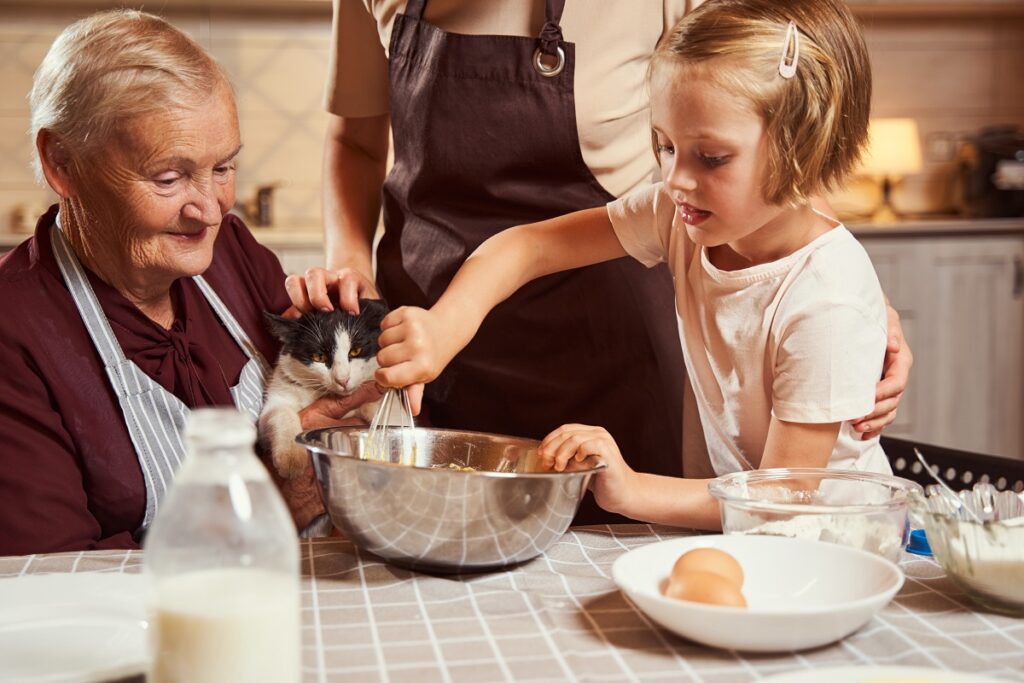 Aged woman and person near her petting a cat and checking how child stirring dough mixture with whisk