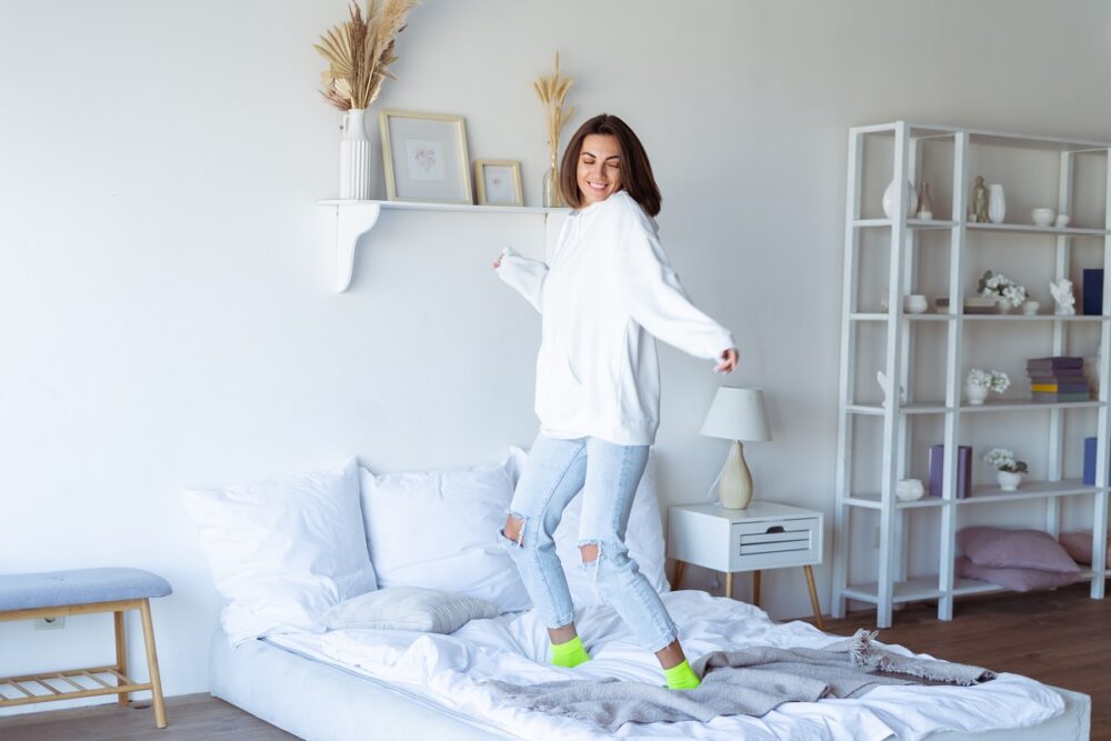 Young woman at home in the bedroom in a warm white hoodie, having fun, jumping on the bed, dancing