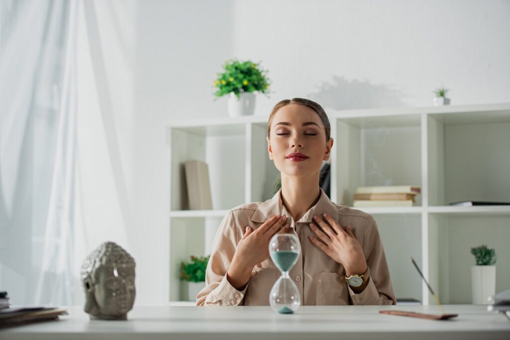 attractive tired businesswoman sitting at workplace with sand clock and buddha head