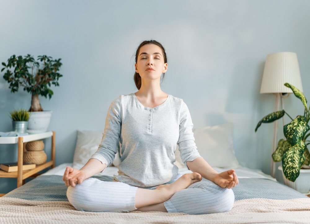 Happy young woman enjoying sunny morning and practicing meditation on the bed.